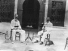 B.Machulka s dalším mužem sedí na židli u malého stolku na dvoře domu, kouří vodní dýmky, Machulkovi leží u nohou pes