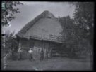 Dřevěná stodola, roubená, příčně průjezdná, dřevěná vrata, zvalbená střecha krytá doškem, před stodolou dvě postavy žen a postava muže, kolem vzrostlé stromy
