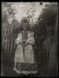 Dívka v partě ve svátečním kroji stojící před dřevěným laťkovým plotem